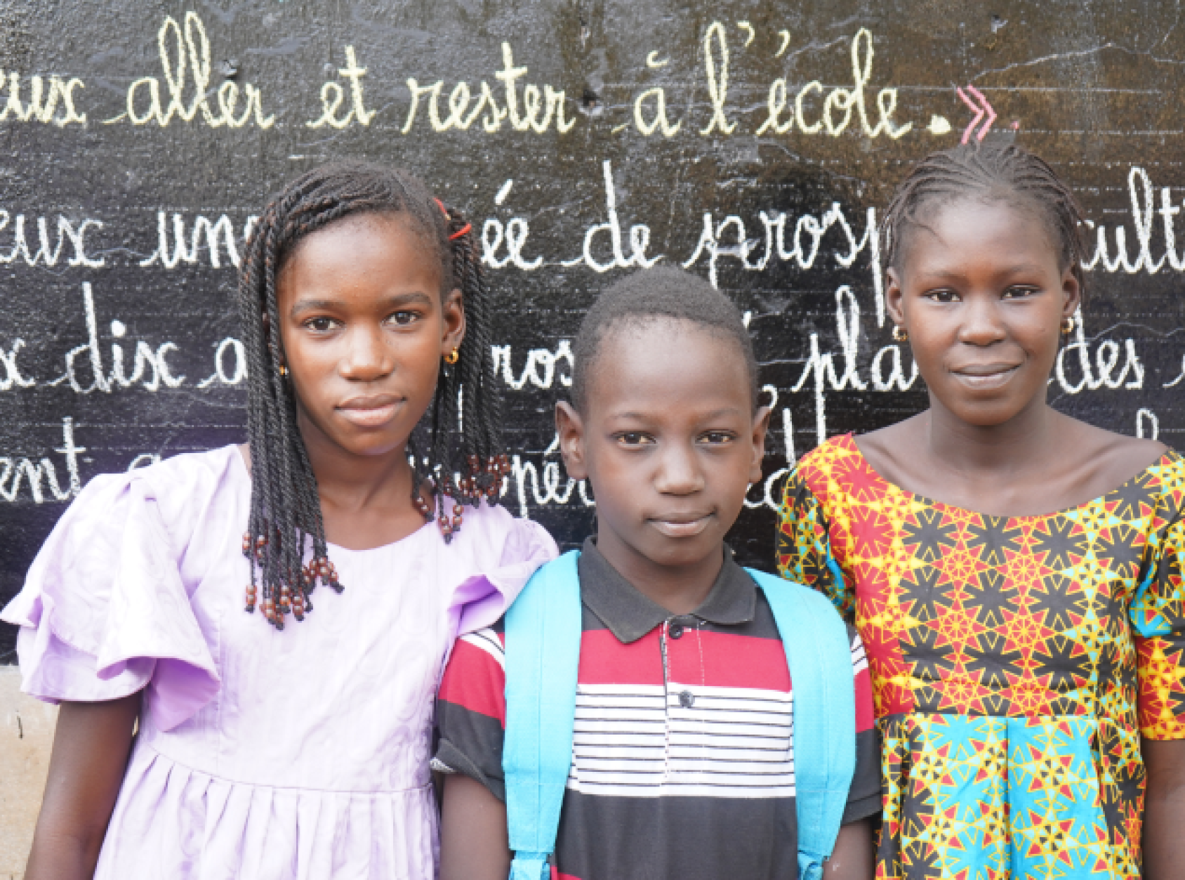 Three children look directly at the camera while standing in front of a chalk board.