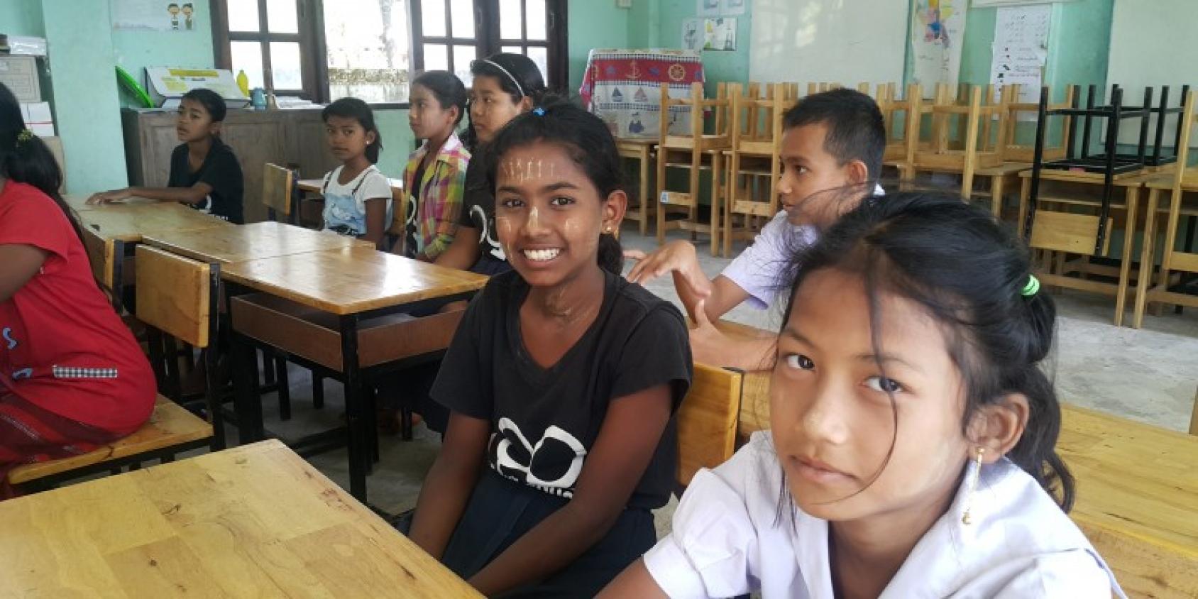 Des élèves thaïlandais sont assis dans une salle de classe. Au premier plan de l’image, deux jeunes filles regardent en direction de l’objectif en souriant. 