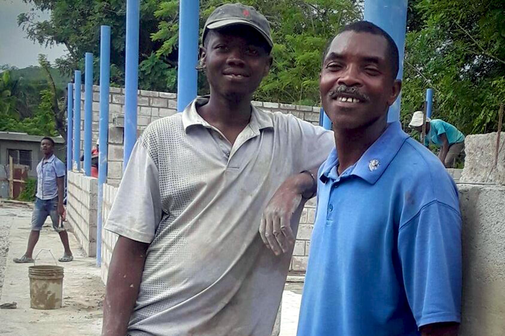 En Haïti, un homme en polo beige et un autre en polo bleu se tiennent debout, côte à côte, devant les poteaux bleus d’une école en reconstruction, et sourient à la camera.