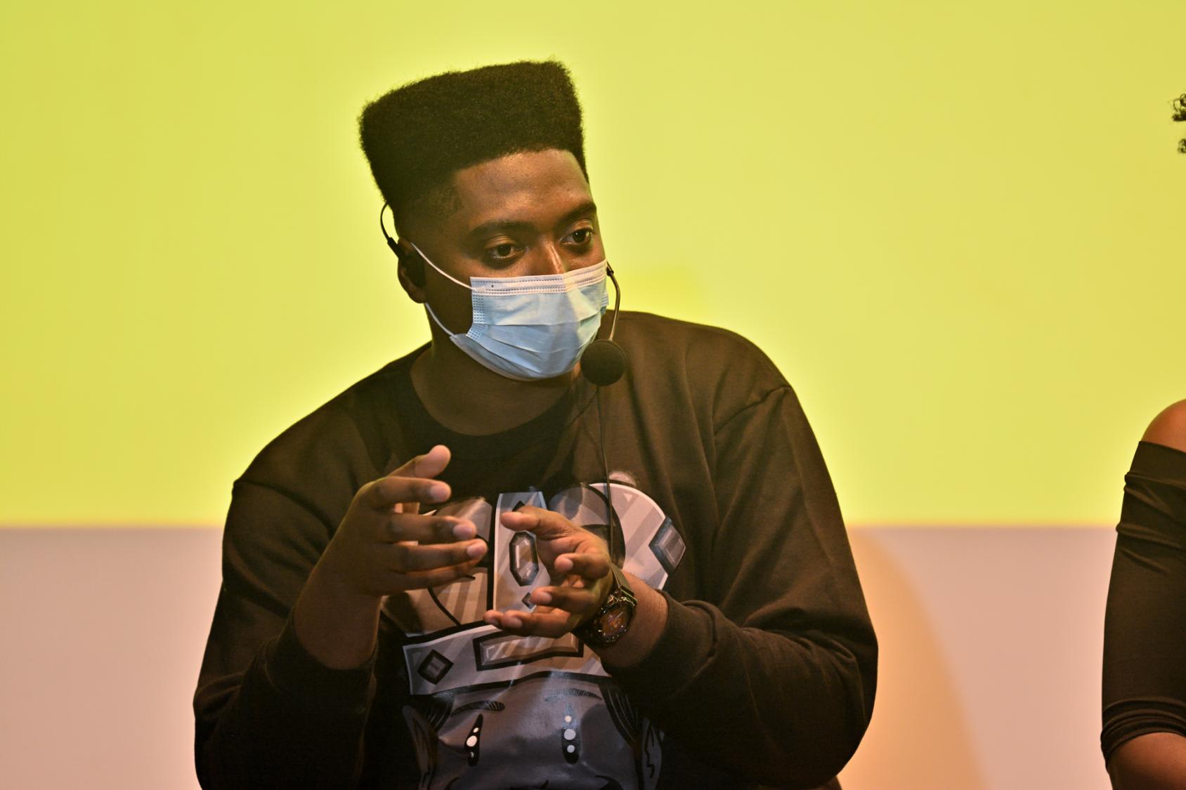 一个戴着面罩的人在一个黄色的投影屏幕前讲话。