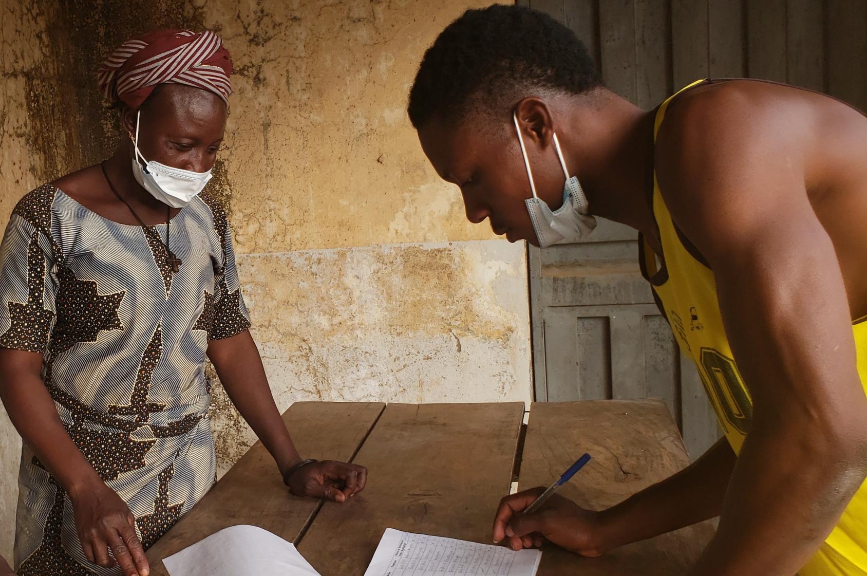 Un homme jeune portant un t-shirt sans manches jaune est penché sur une table en bois et écrit sur une feuille de papier. Une femme se tient de l'autre côté de la table.