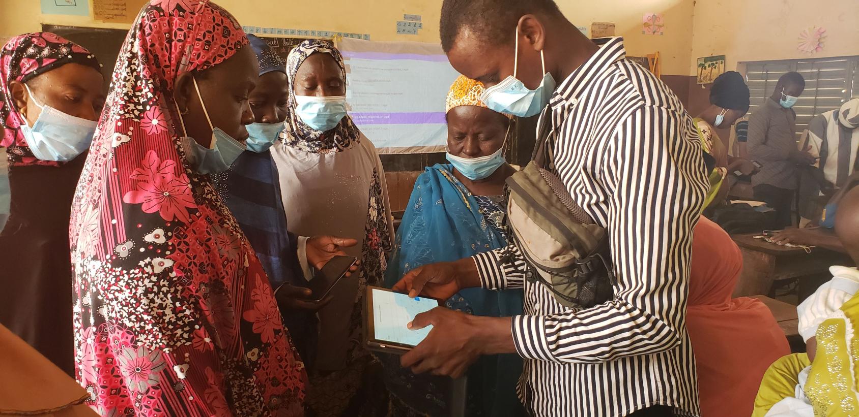 Un jeune homme portant un masque de protection respiratoire se tient debout, entouré de plusieurs femmes, elles aussi masquées. Il tient une tablette à la main et leur montre comment l'utiliser.