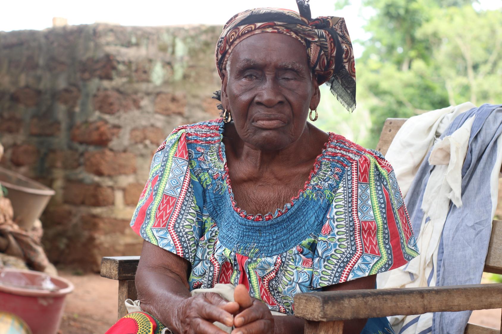  Une femme âgée vêtue d'une robe aux multiples couleurs et les cheveux attachés à l'aide d'un foulard, est assise sur un banc en bois et regarde en direction de l'objectif.