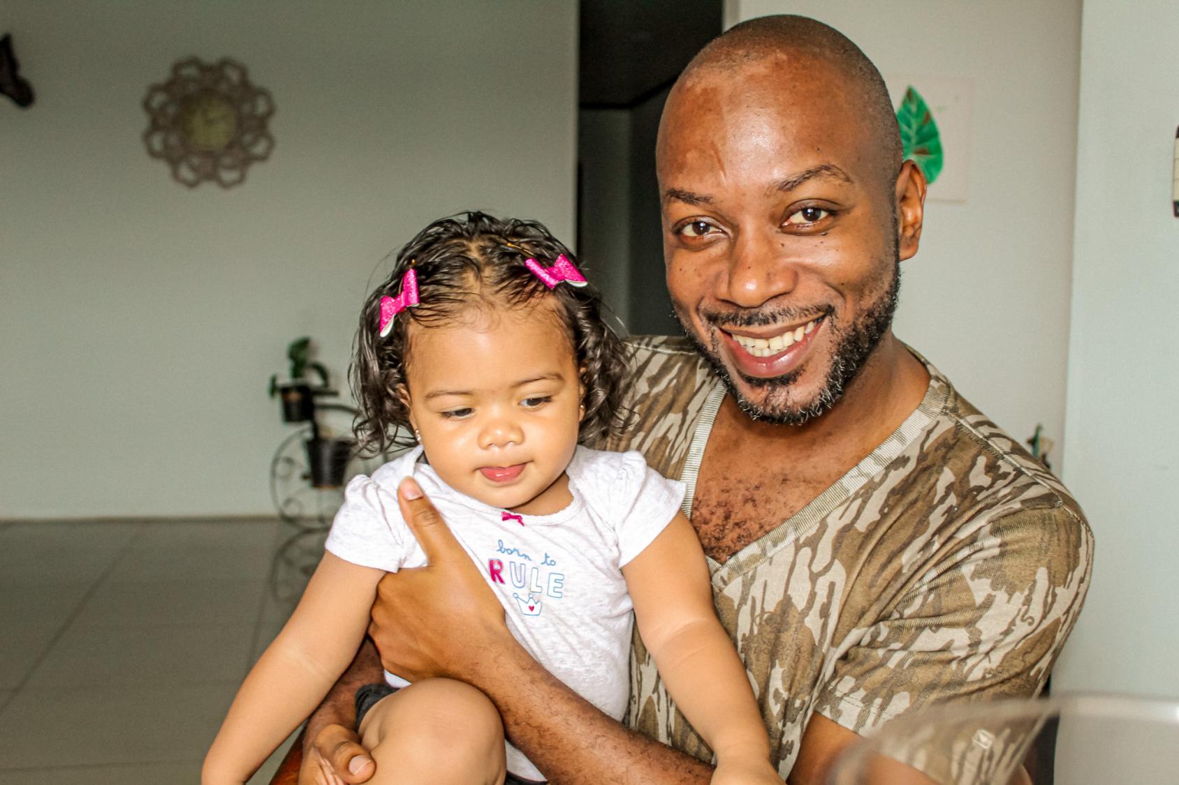 A man sits in a room and holds a child with pink barettes on his lap.