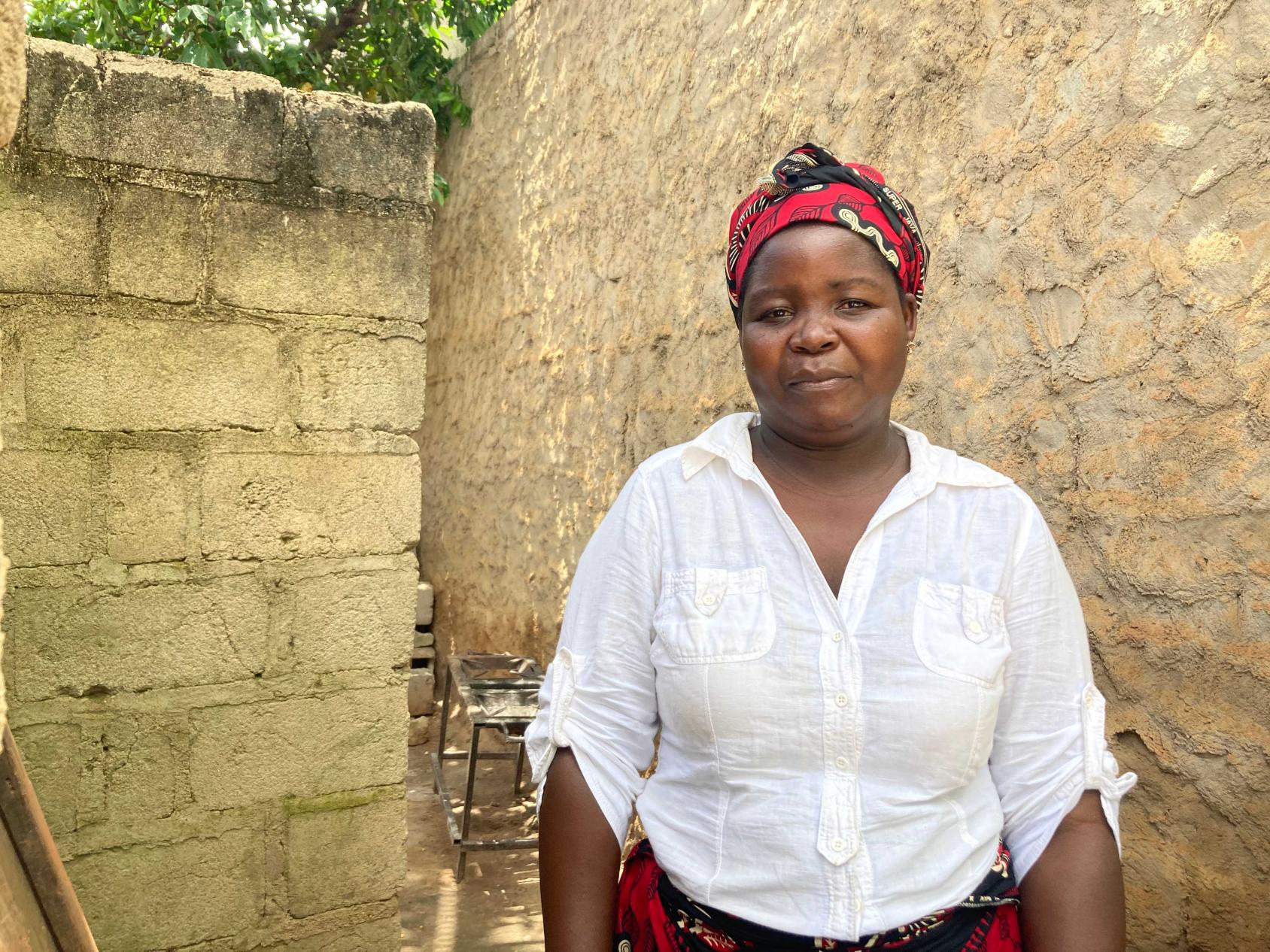 امرأة ترتدي قميصًا أبيض وتنورة، تبتسم للكاميرا بينما تقف أمام جدار حجري.