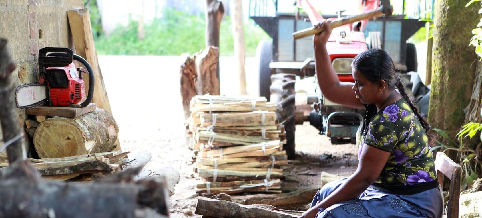 A woman sits on the ground and hammers wood into smaller kindling.