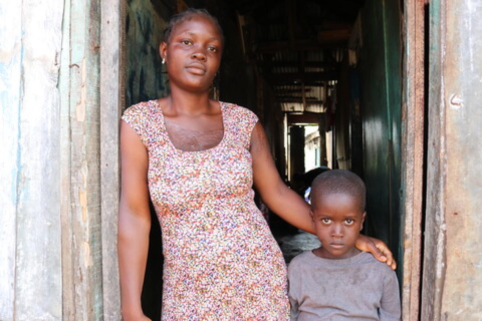 一位年轻的利比里亚妇女站在自家门口，她的手臂搂着她的小儿子的肩膀。