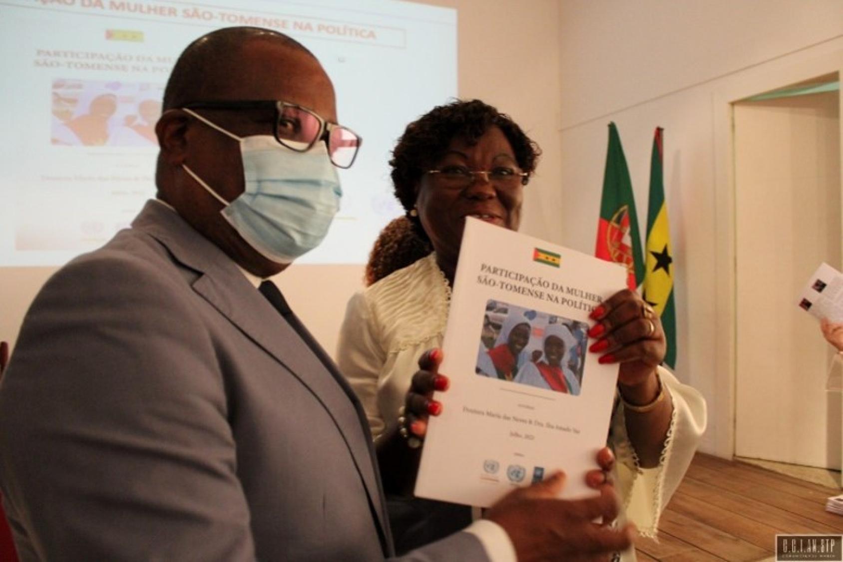 Un hombre, con traje y mascarilla, sostiene un manual que le ha entregado una mujer. Hay banderas de Santo Tomé y Príncipe en el fondo.