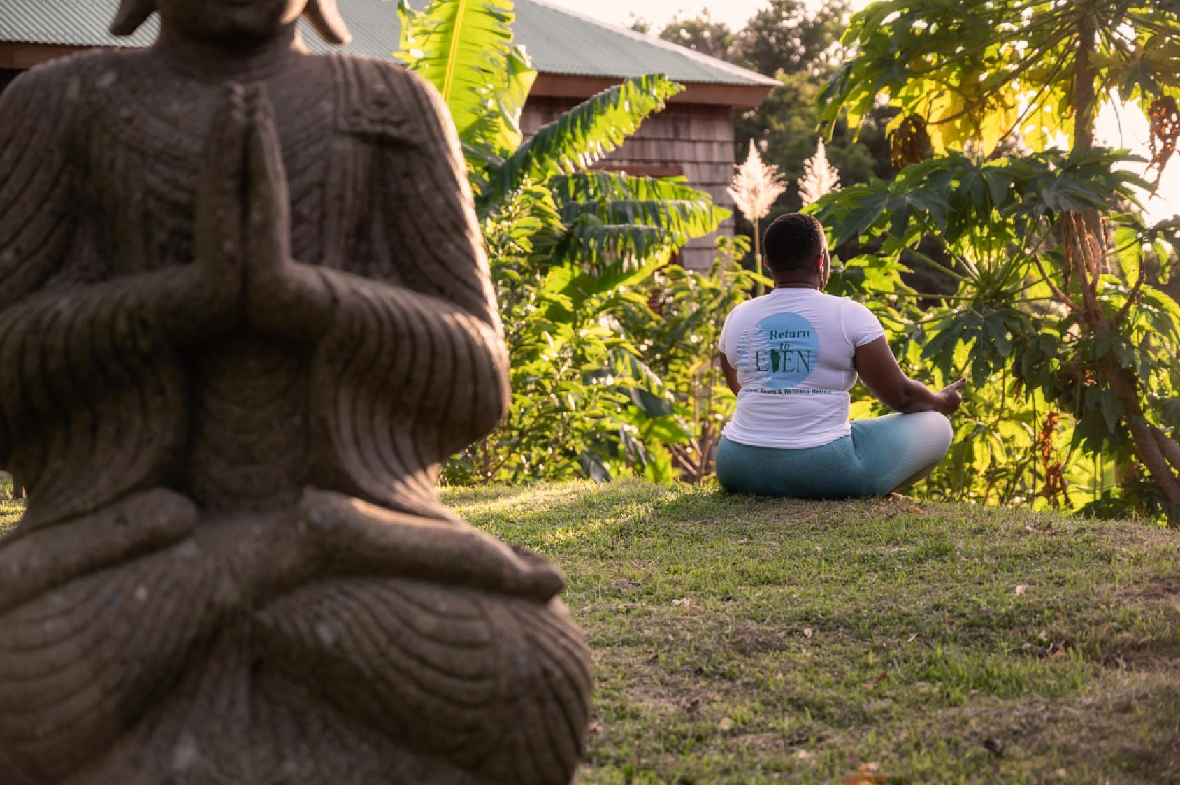 Une femme aux cheveux courts portant un jean et un t-shirt blanc est assise en tailleur, dans l'herbe, les mains posés sur les genoux, devant une végétation luxuriante, et médite. Au premier plan de l’image se trouve une statue représentant un personnage également en position de méditation.