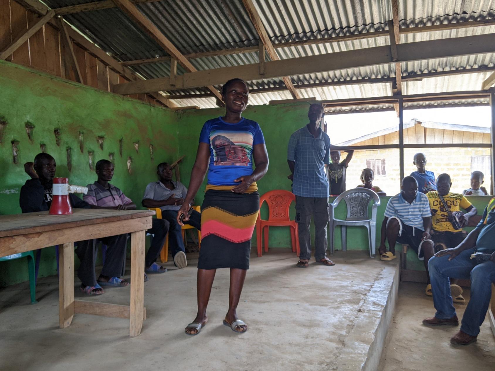 In Liberia, a woman stands on a stage, surrounded by several people sitting on chairs in a room with a roof made of wood and metal sheets.