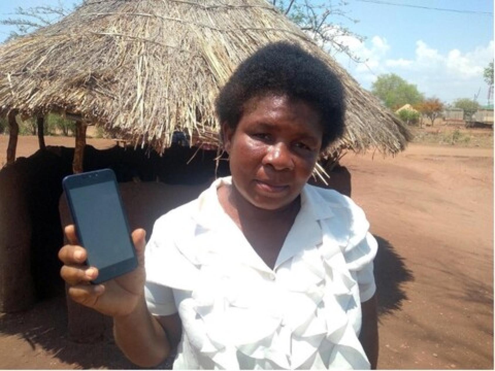A woman in a white shirt stands in front of a hut-like structure, and holds her cel phone in front of the camera.