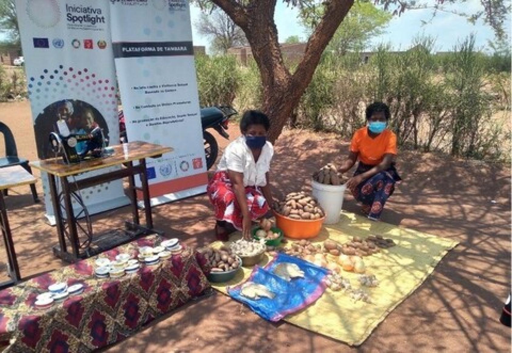 Two women sit outside in tan field under a tree, with signs from Spotlight Initiative behind them.