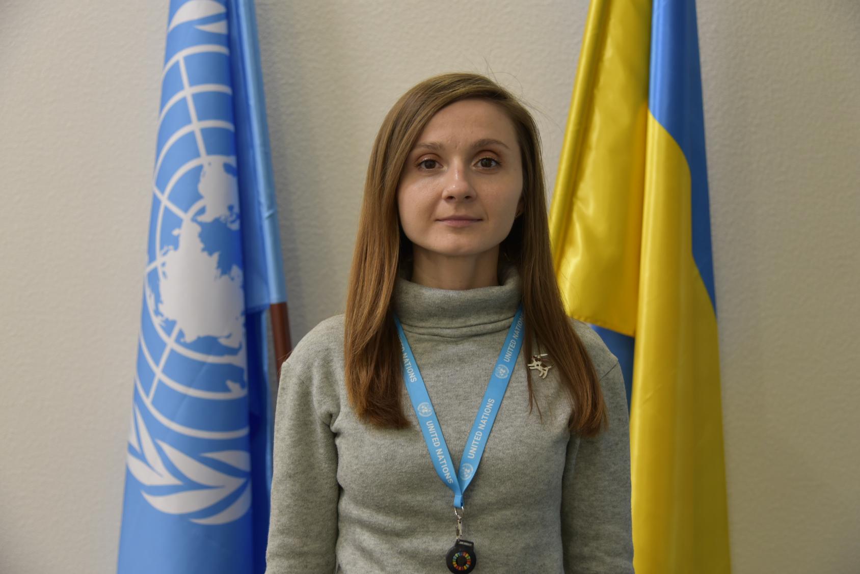 A woman, a UN team member, stands in front of a flag of the UN and another flag of Ukraine.