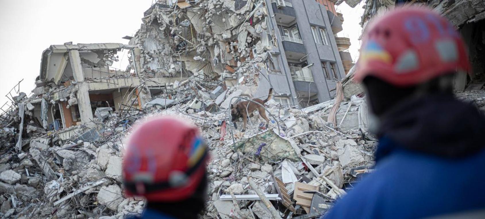 Figures in metal hats stand before debris from buildings in the aftermath of an earthquake.