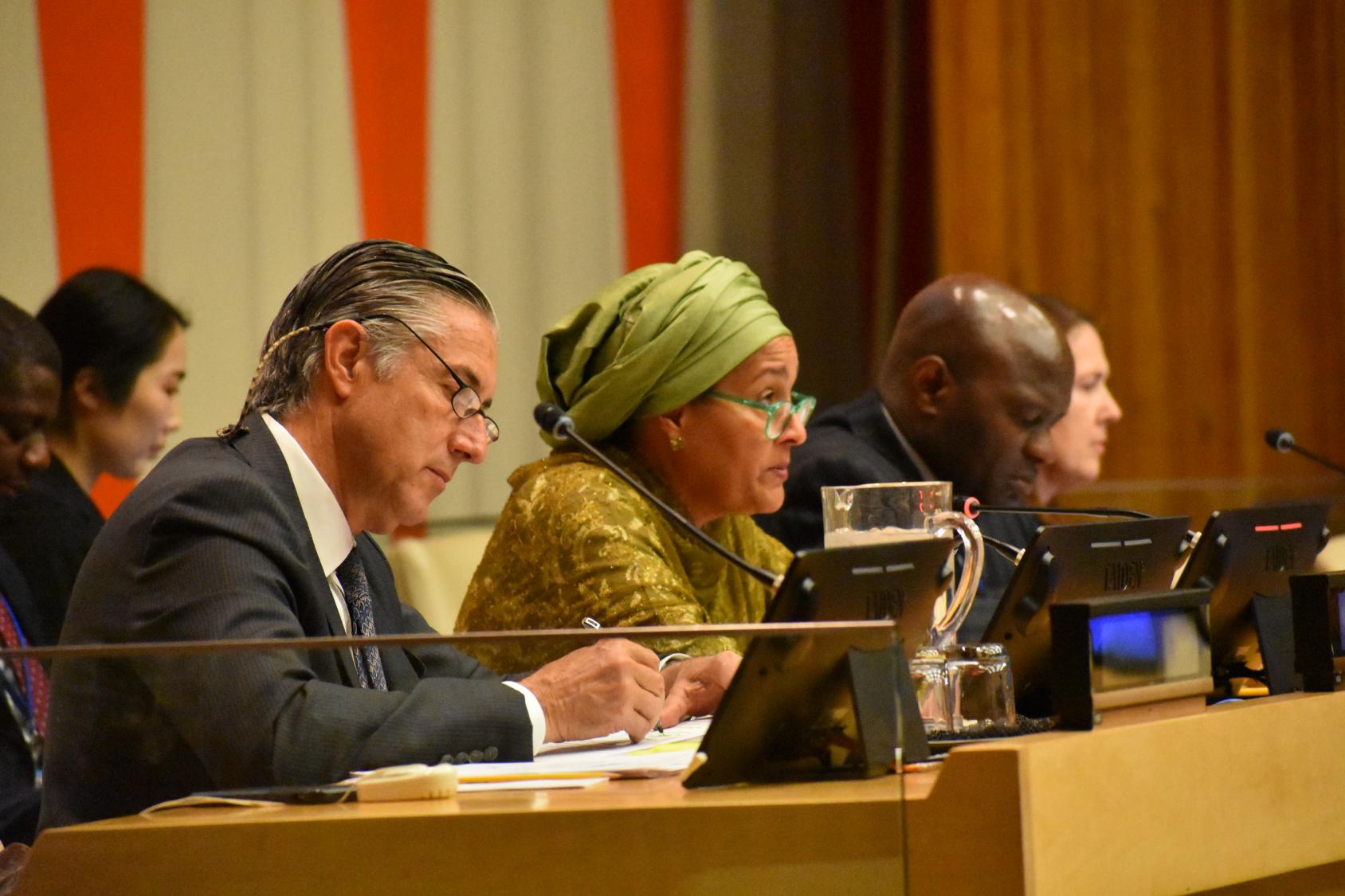Man in a black suit and a woman in a green dress sit at a podium and speak into a microphone