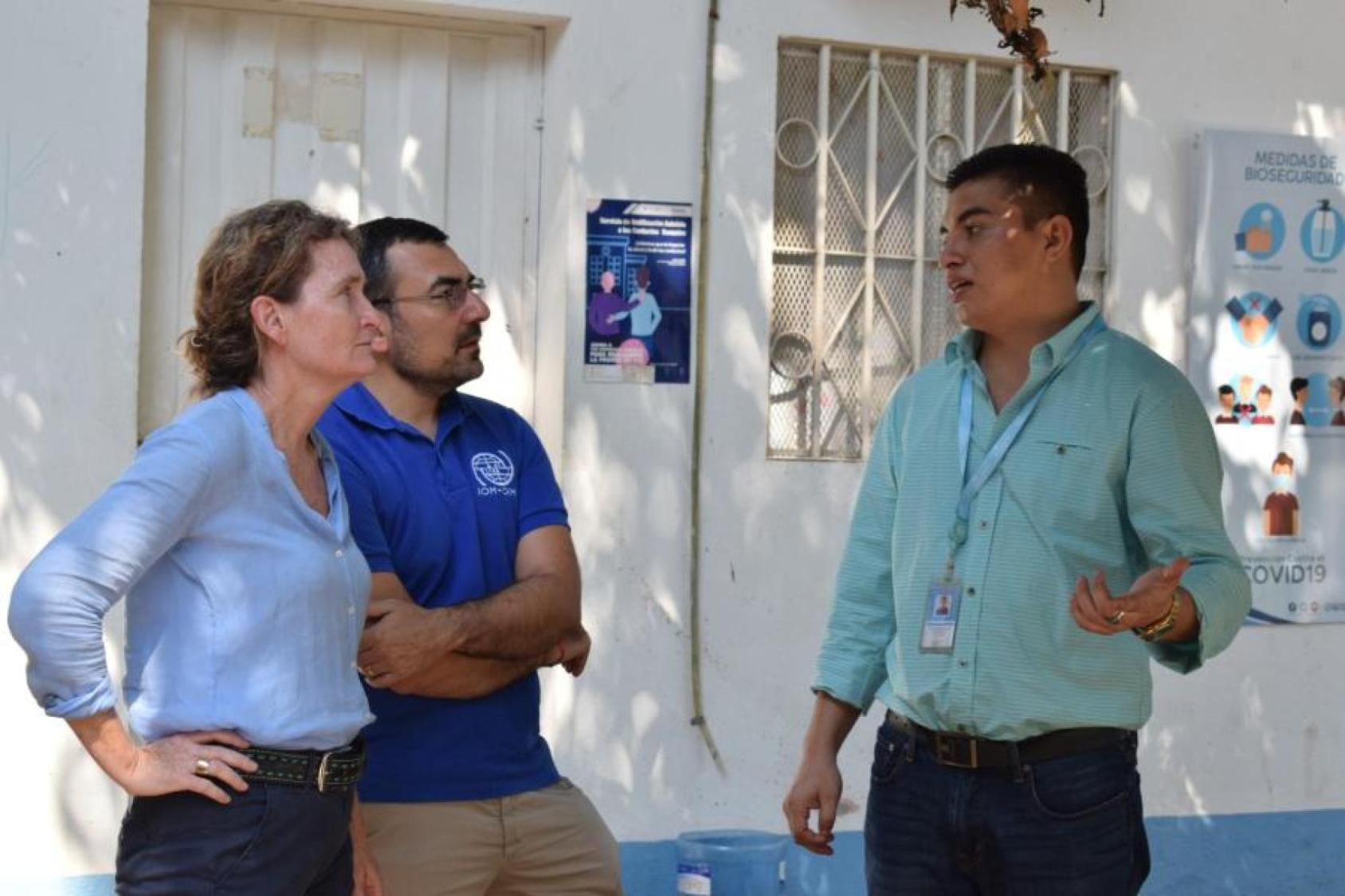 A woman in a blue shirt and a man in a blue shirt and spectacles listen to another man in a green shirt talking animatedly