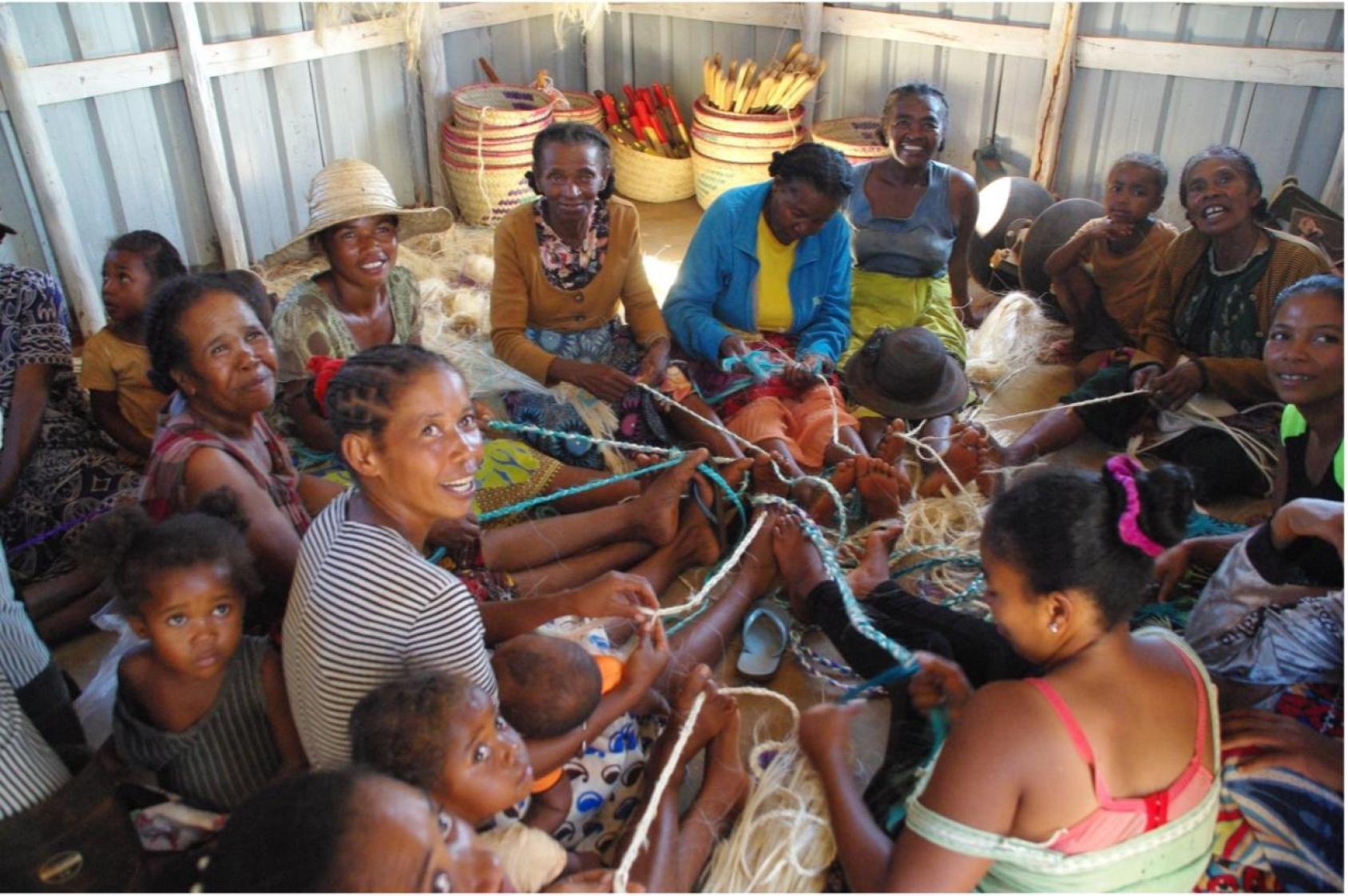 Group of people in multicoloured clothes sitting in a hut in a close circle passing woven threads between them
