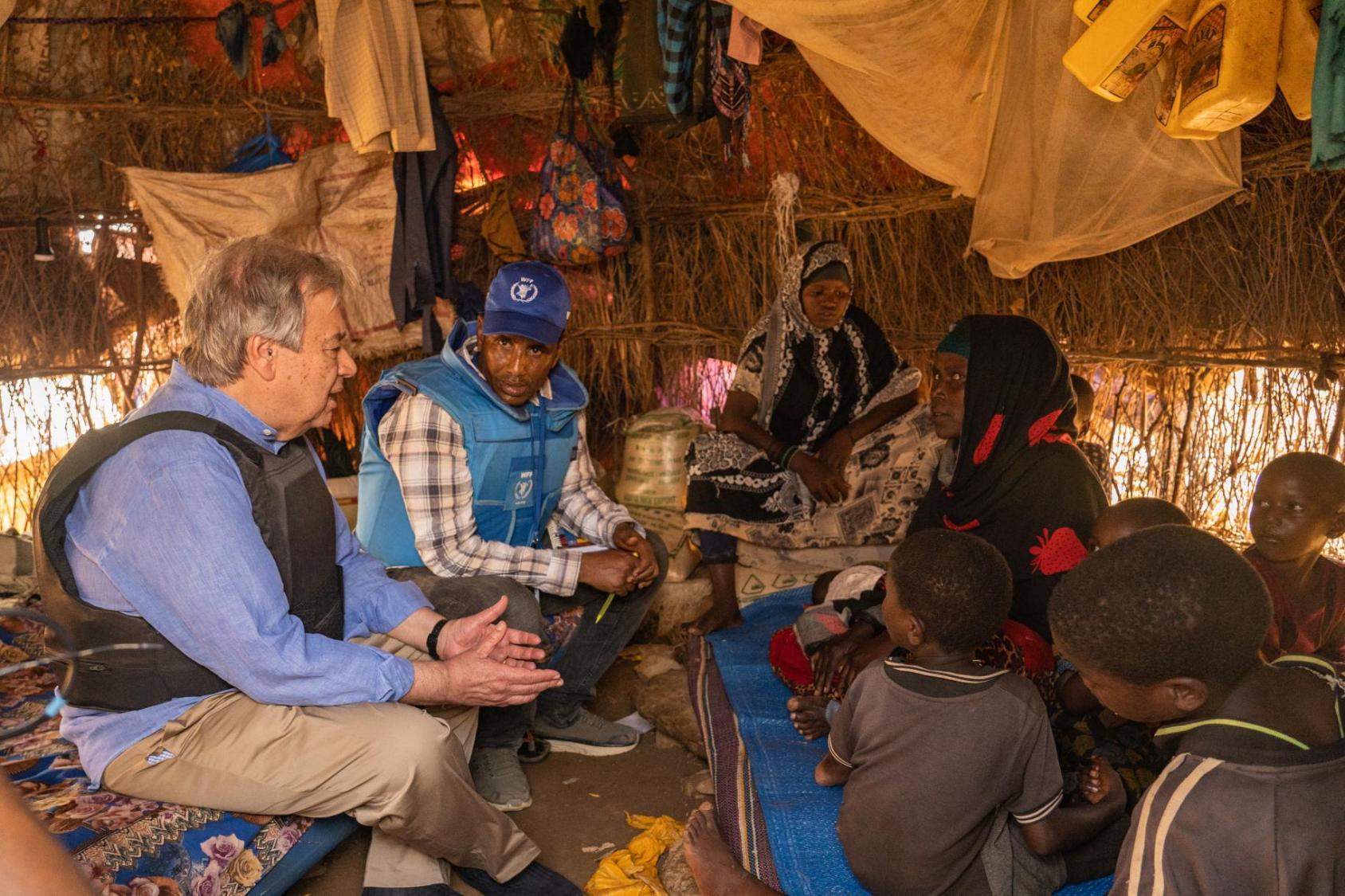 A man with grey hair and a blue shirt with a vest sits in a tent talking with people in blue vests and in other colourful clothes