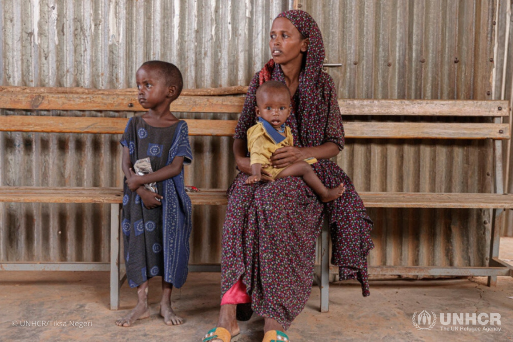 A woman in a brown dress holds a baby on her lap as a small child dressed in blue sits to her right on a wooden bench