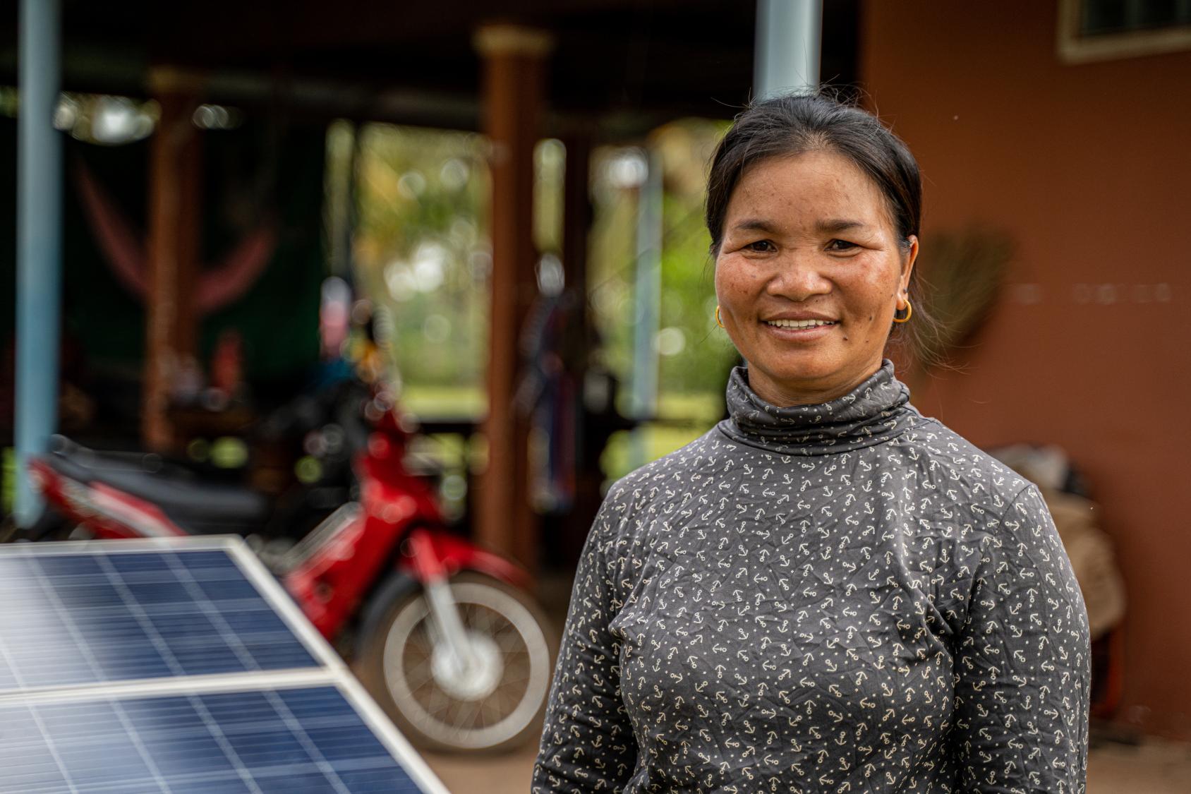 A woman in a grey shirt stands in front of a solar panel in rural Cambodia