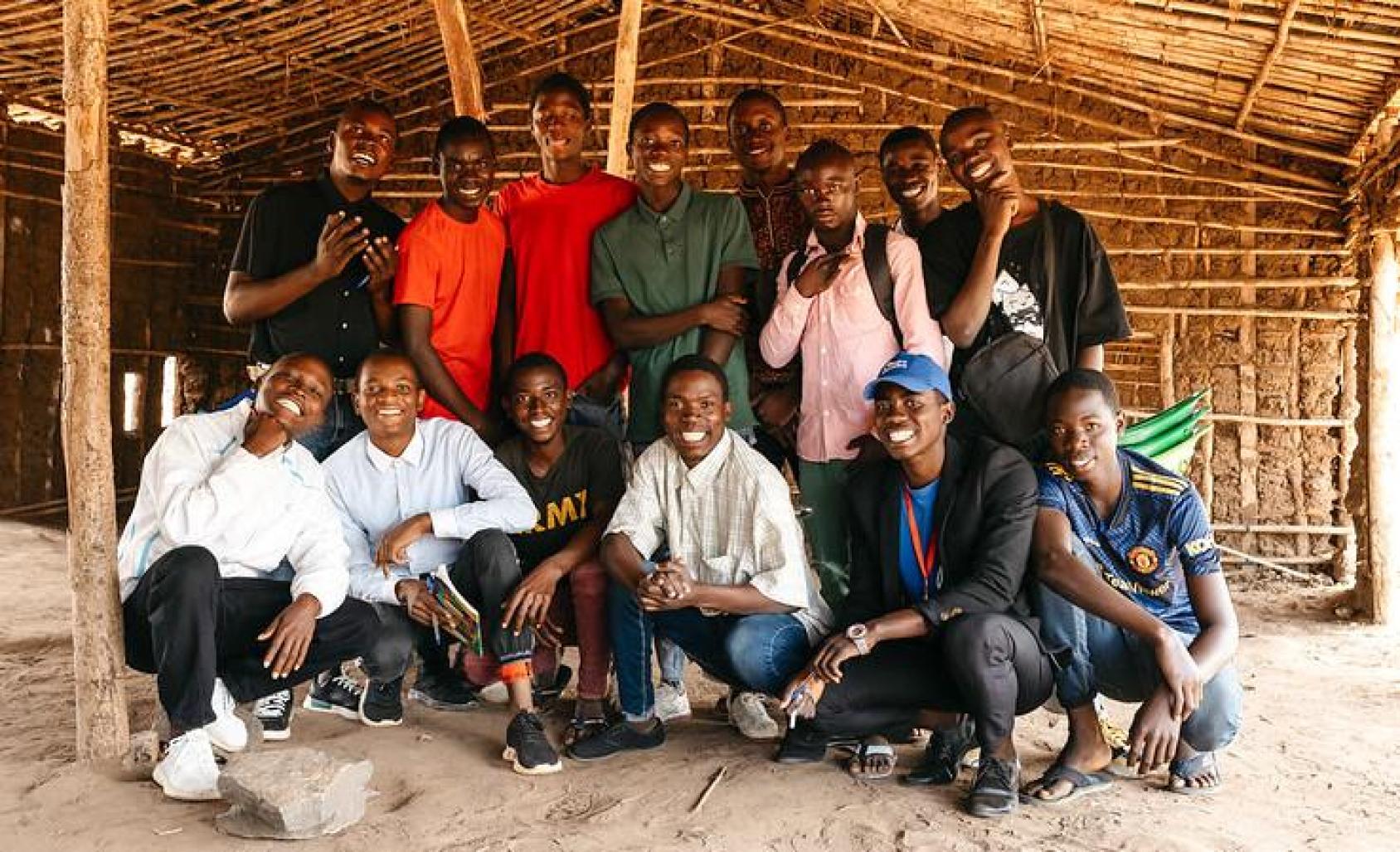 Fourteen young boys in colourful shirts and pants in a bamboo shed