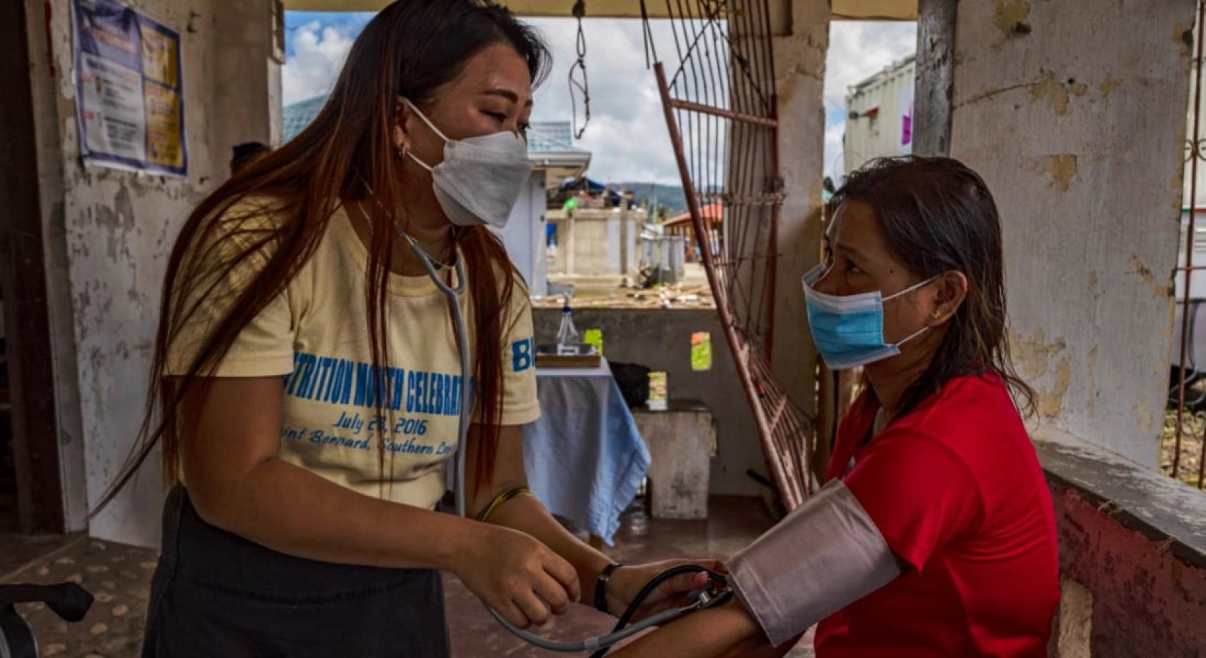 A medical professional in a mask checks the blood pressure of a woman in a red shirt as she sits amidst the debris