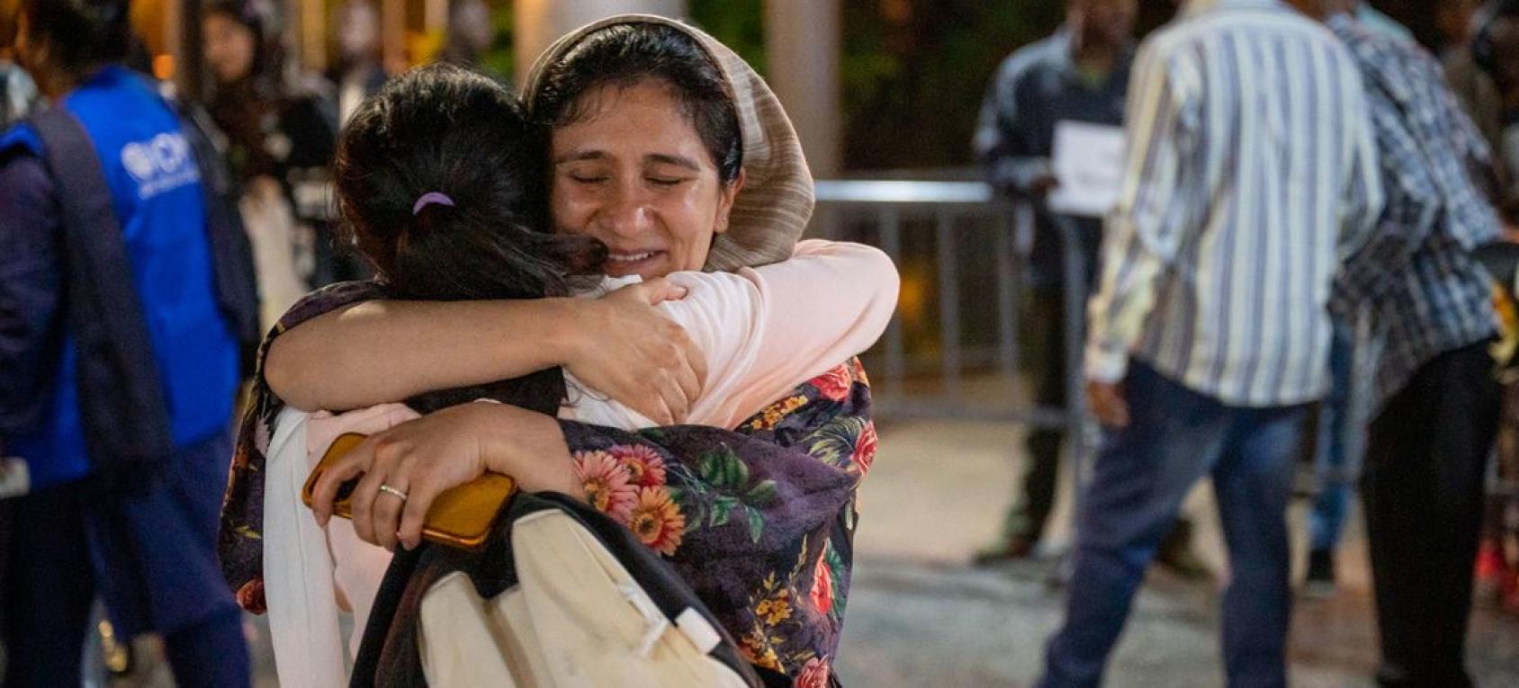 Two women embrace, and one is wearing a hat and holding a phone in her hand.