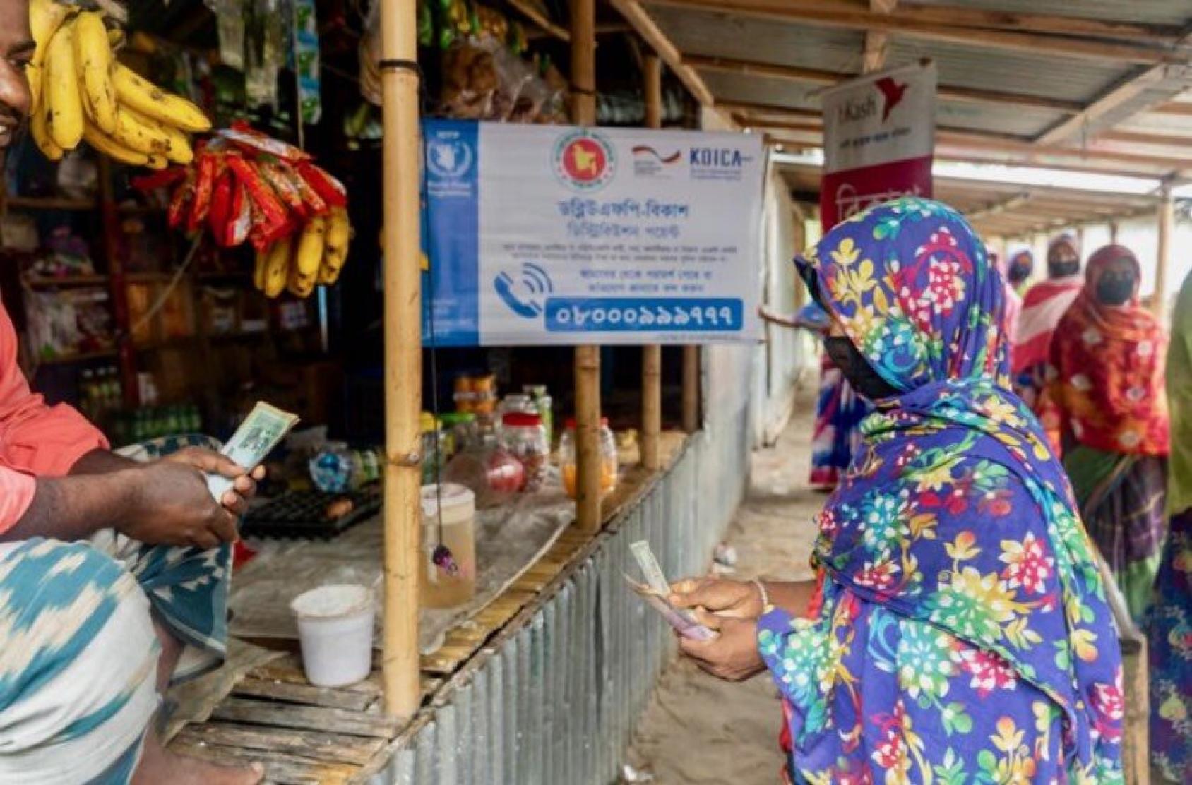 A woman in a colourful saree holds out some money to a vendor in a market where different items including bananas hang.