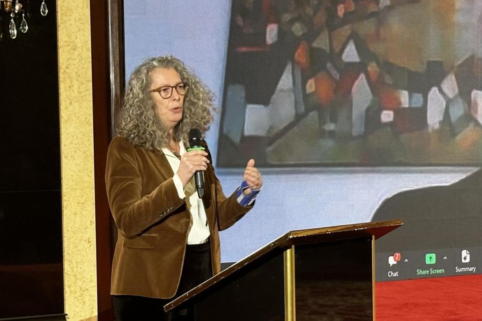 A woman in a brown coat with curly grey hair and glasses speaks into a microphone at a podium