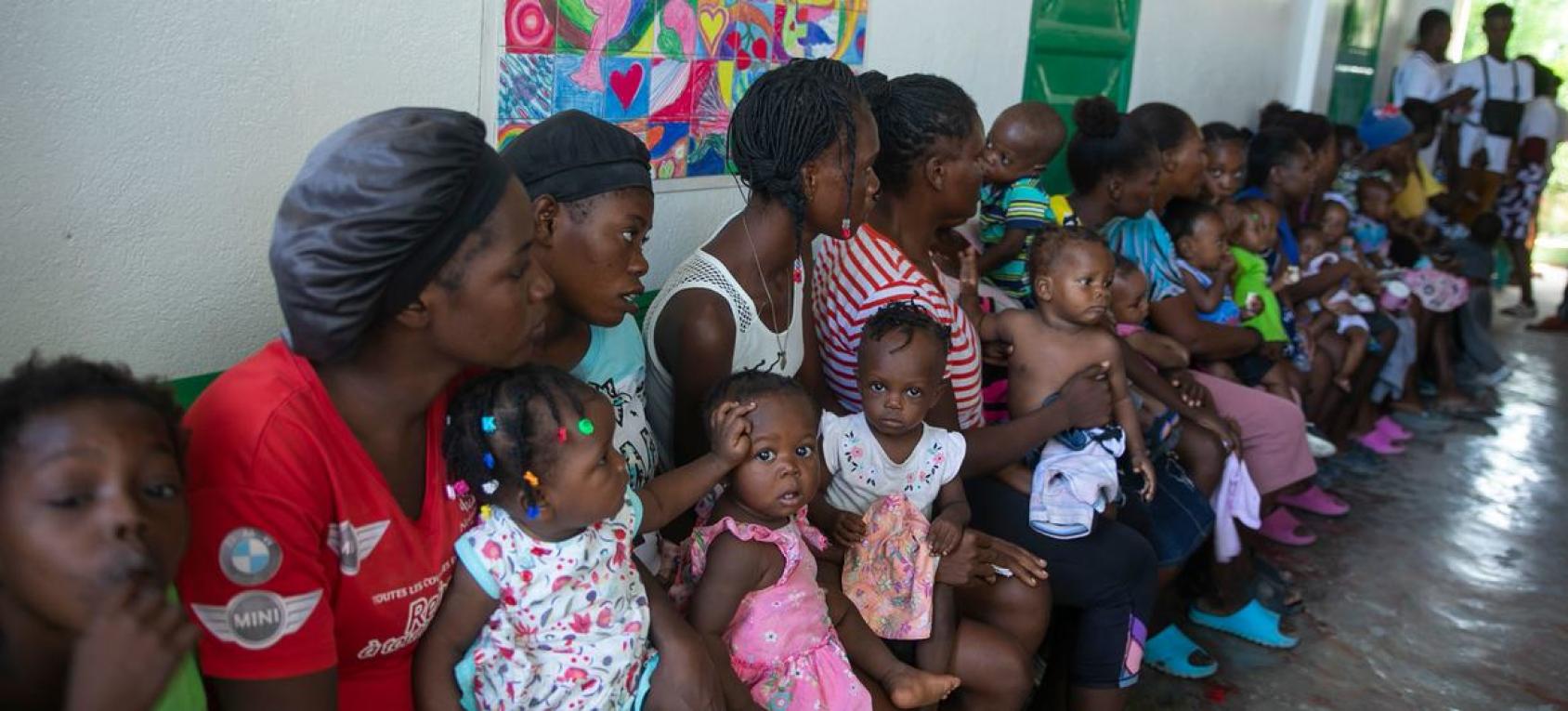 A row of women and children, all in multicoloured clothes, lined up against a wall. The children are sitting on the laps of the women.