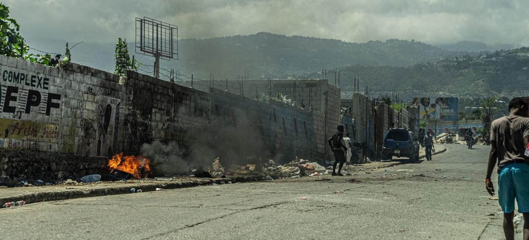 A scene of a road engulfed in grey smoke while fires burn in the distance and vehicles are parked.