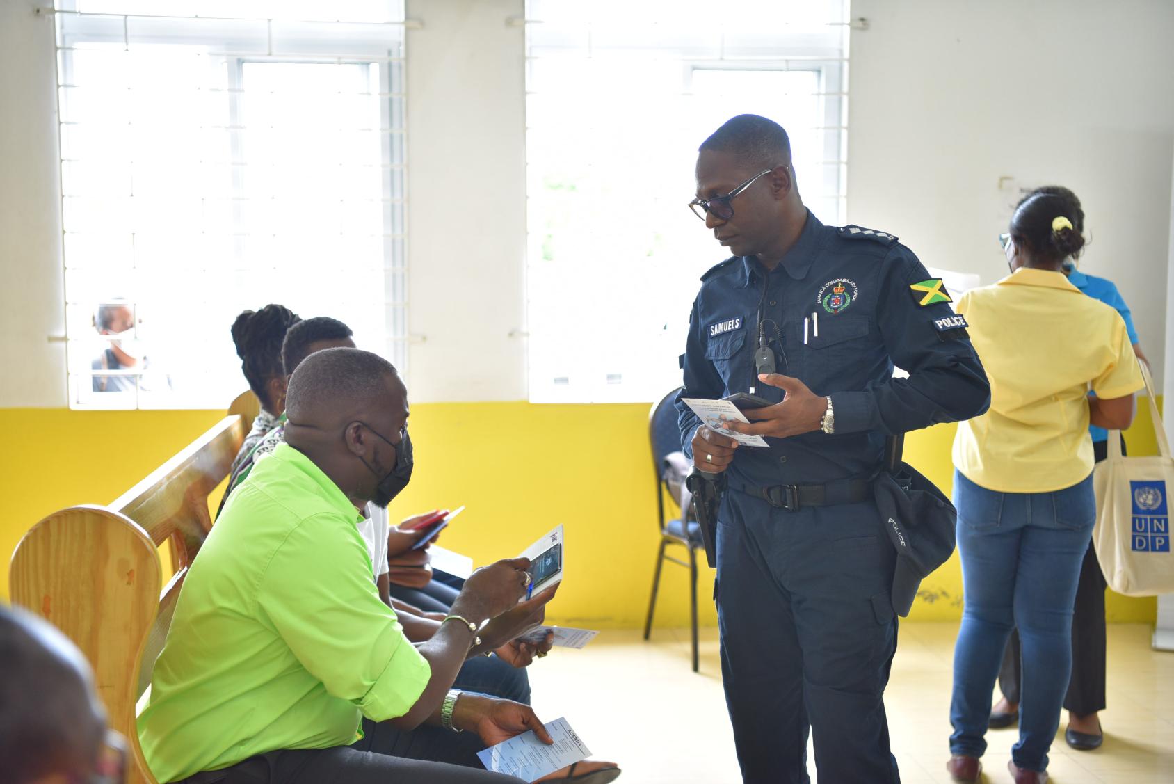 A policeman in dark blue clothes talks to people sitting on chairs in yellow and green shirts