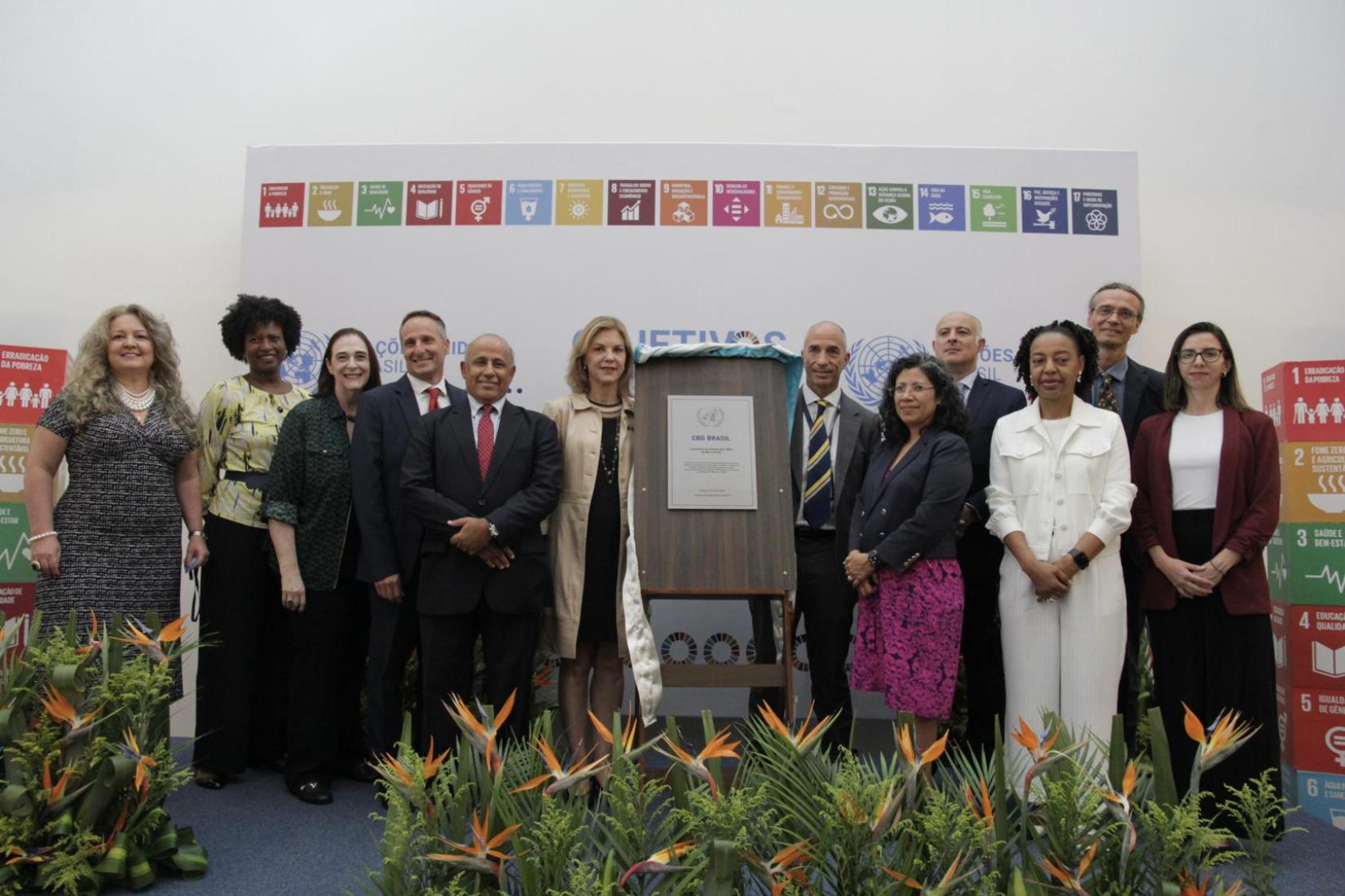 A group of formally dressed individuals stands in front of a backdrop adorned with the SDGs.  Ornamental plants with orange flowers decorate the foreground