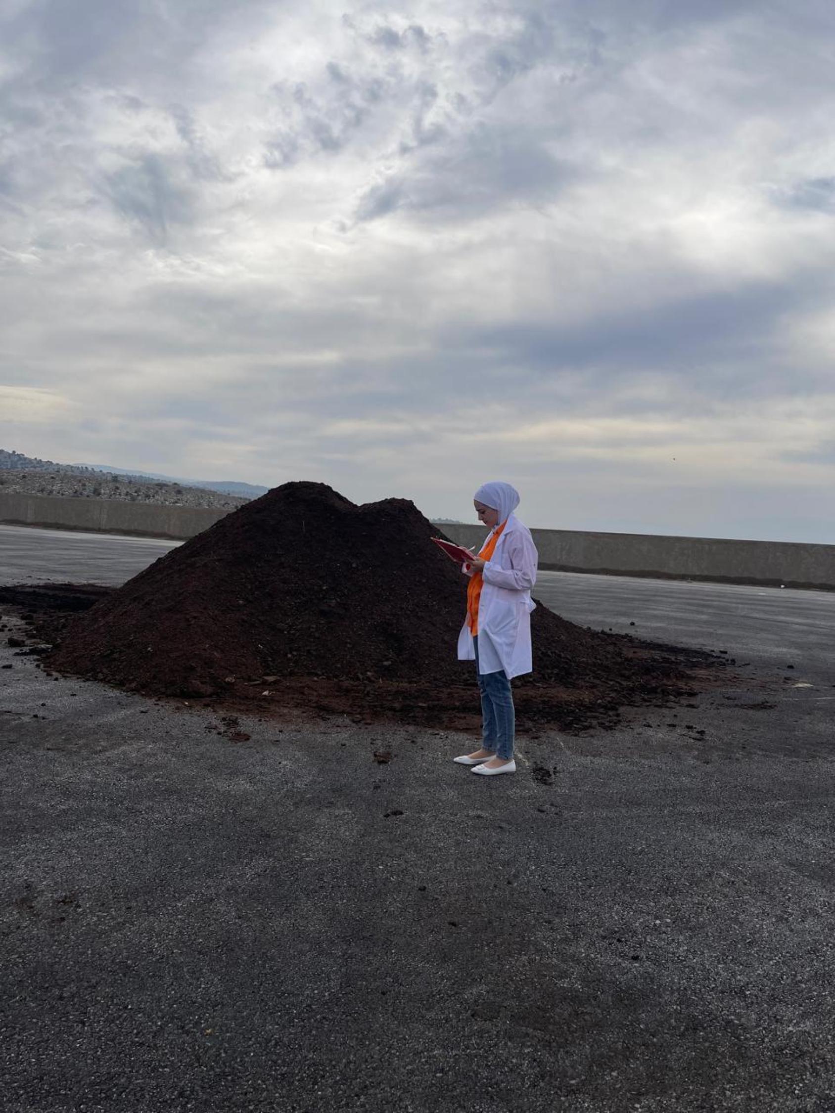 A woman in a white dress stands in front of a mound of black soil fertilizer