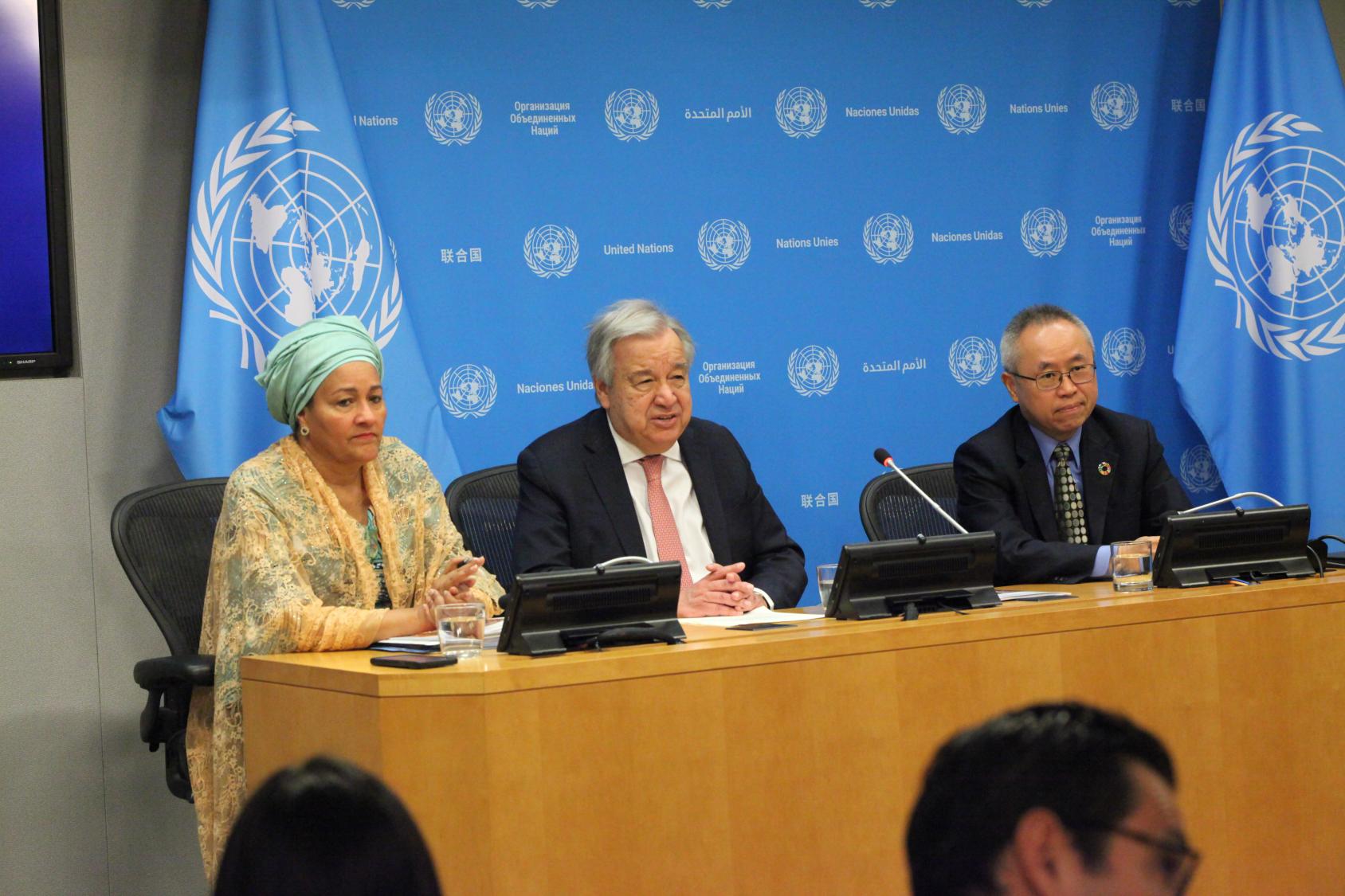 A woman and two men seated at a conference room table. UN flags besides them and a UN backdrop behind them