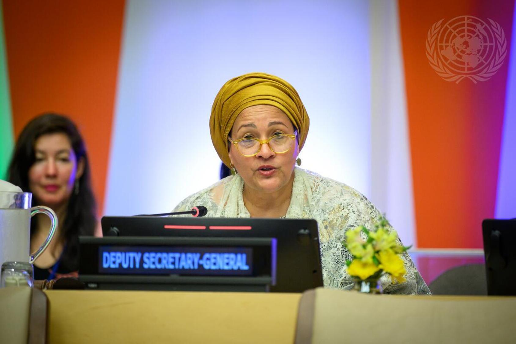 A woman in a yellow headscarf and spectacles speaks into a microphone at a podium