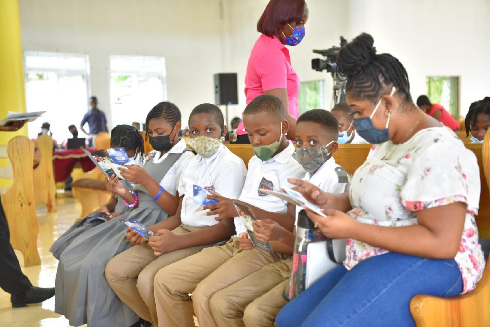 A group of students in school uniforms and masks lined up next to a teacher, an older woman in a blue skirt holding an electronic tablet
