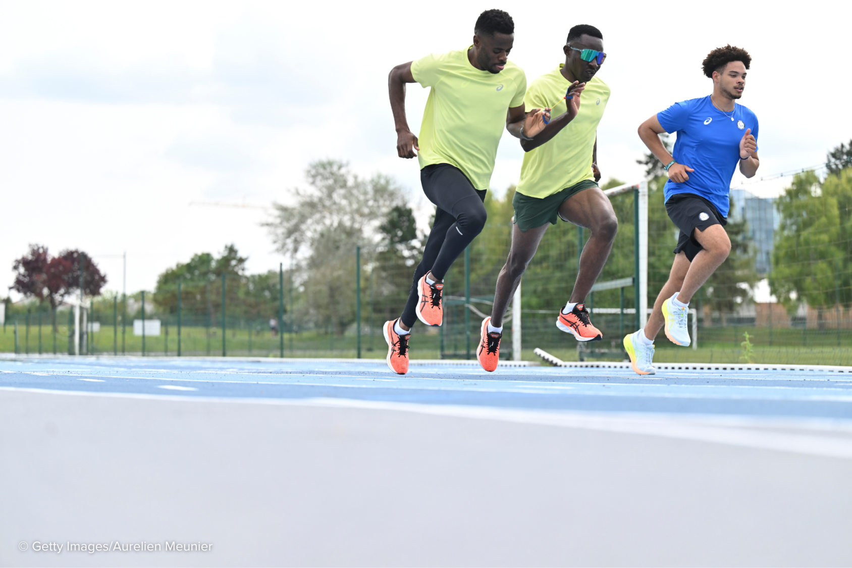 Three men, two in a fluorescent shirt and one in a blue shirt run on a blue race track. The man in the middle 