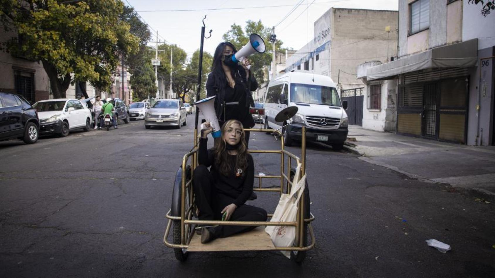 Two women wearing black clothes with bullhorns are traveling in a makeshift vehicle through the streets on Mexico