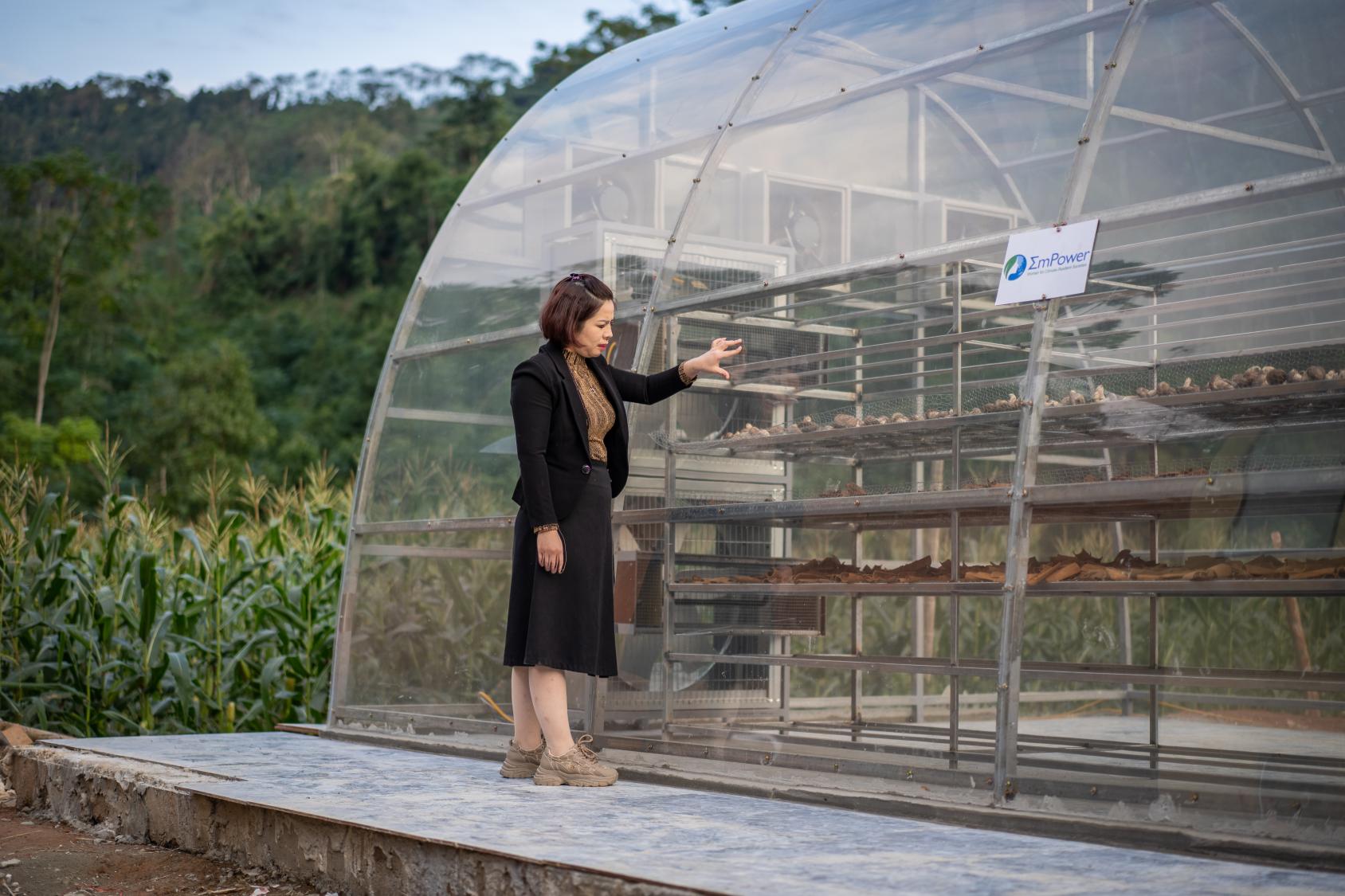 A glass house with plants inside it and a woman in a black suit stands right outside.