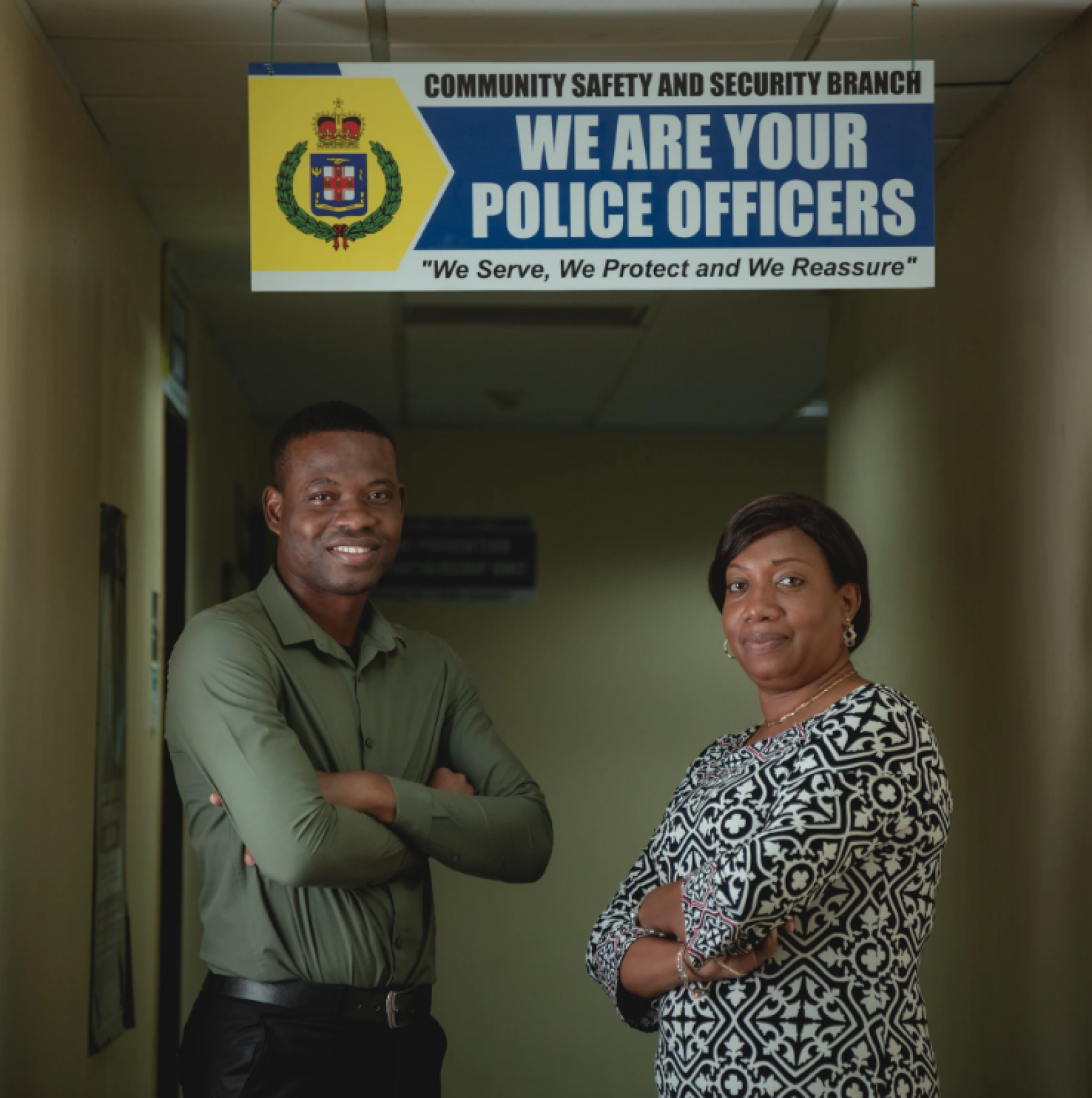 Two people, a man and a woman, in a dark shirts and blouses stand cross-armed underneath a board that that reads "we are your police officers" along with a symbol.