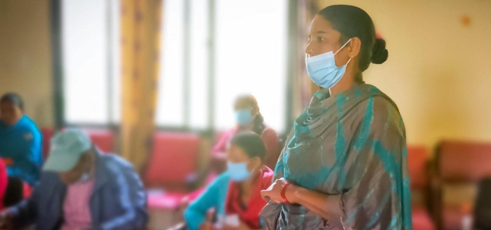 A woman in a green saree and a mask covering her mouth stands in a room while other women are seated in the background