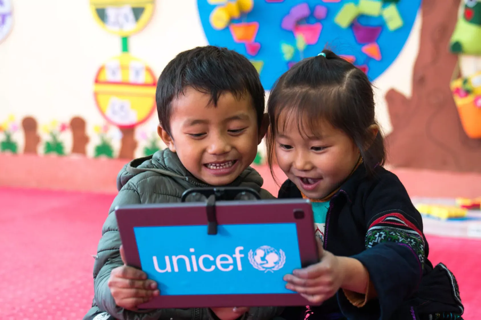 Two children look at an ipad screen with glee. A UNICEF logo appears behind the device.