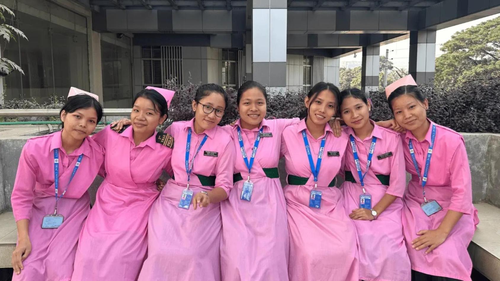 A group of seven young women in pink nurse uniforms sit next to each other outside a hospital building