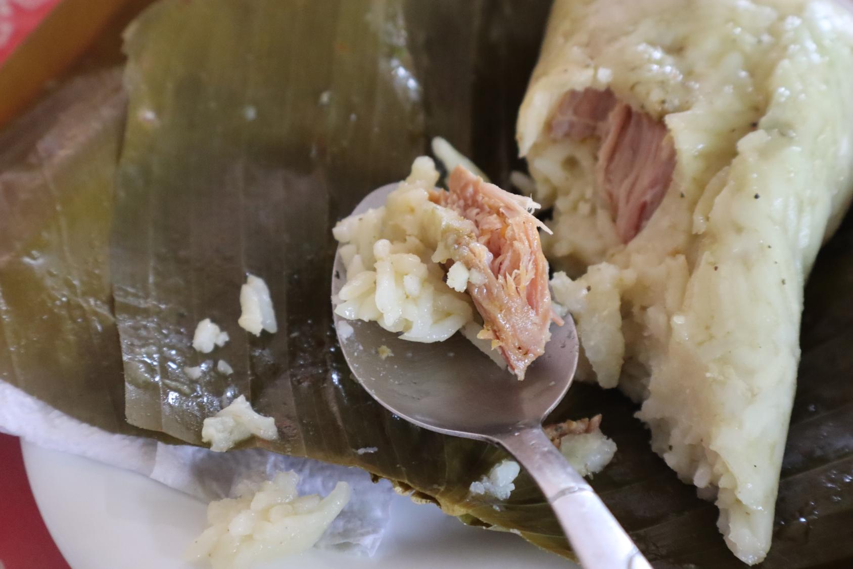 A close-up picture of a spoon with rice and meat in a corn casing, wrapped in a dried banana leaf.