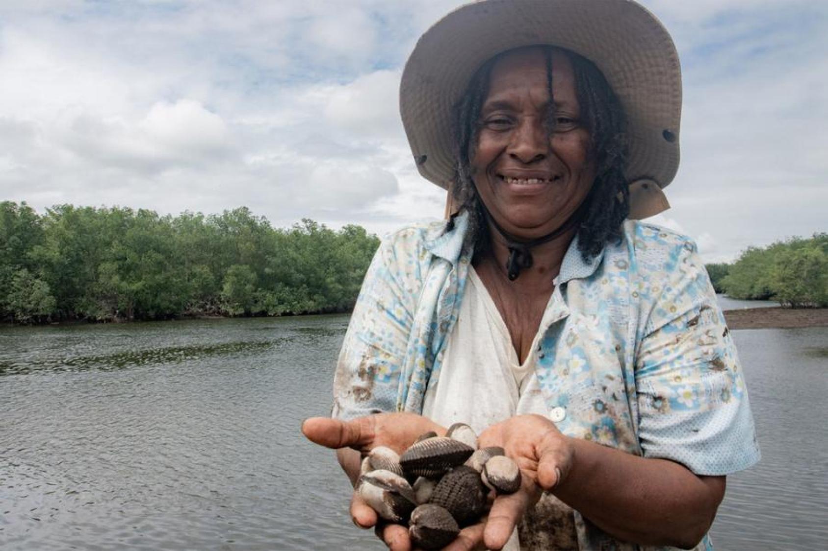 A woman in a white shirt and a hat holds up her hands that carry a bunch of tiny mollusks. In the background is a river with lush green foliage.