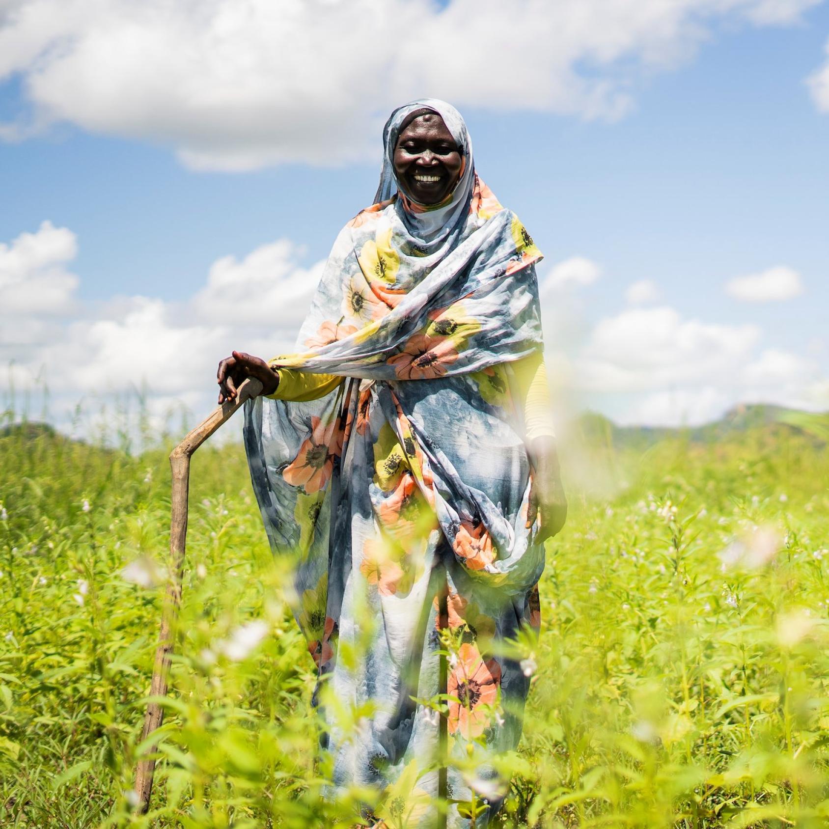 A woman in a blue dress with a headscarf makes her way through a green farm with a sickle-like tool.
