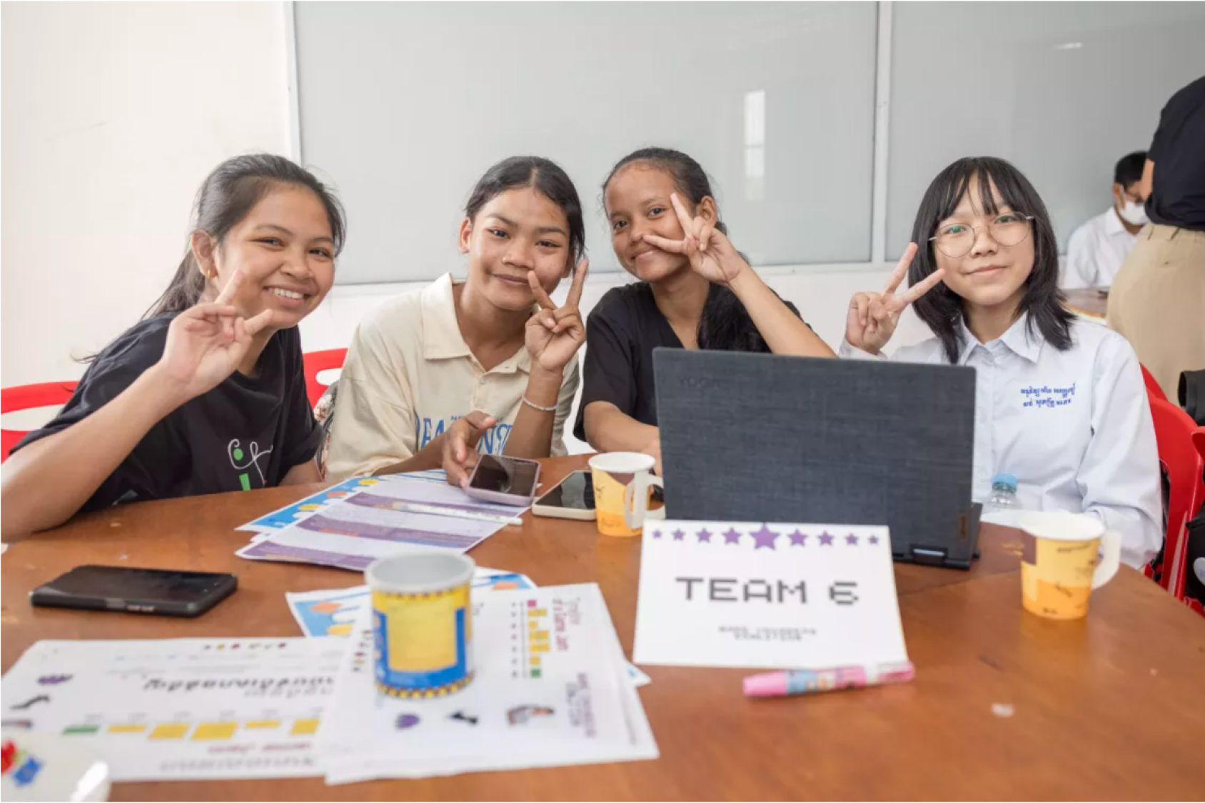 Four school girls in uniform sit around a table with computers, smiling at the camera with their fingers raised in a peace sign.
