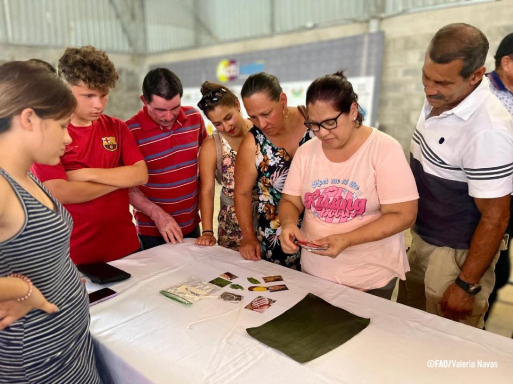A group of people in colourful clothes gather around a woman dressed in a pink shirt who appears to be reading a card.