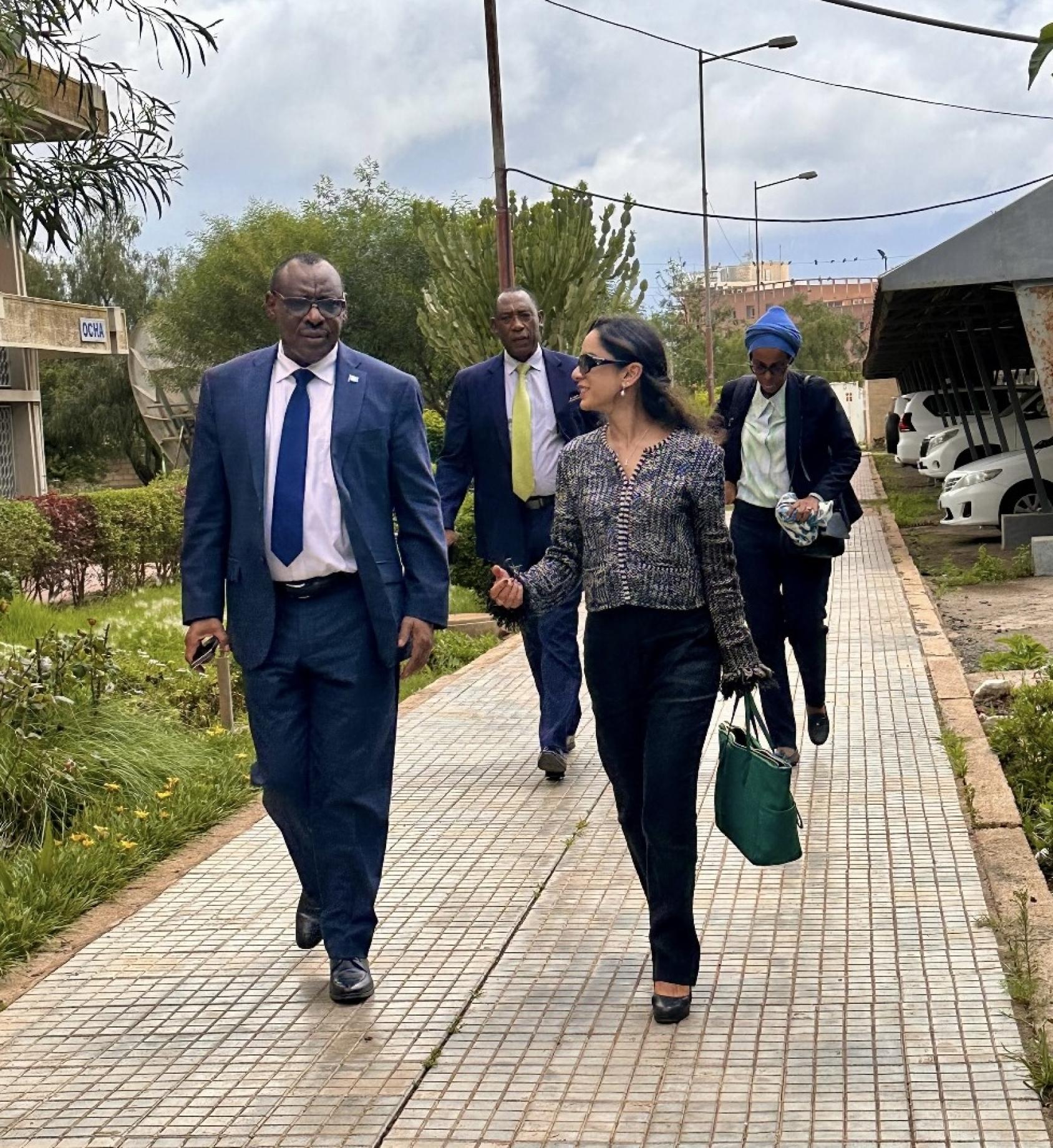A woman in a black formal dress and sunglasses walks and talks to a man in a navy suit. They are followed by two men in suits.